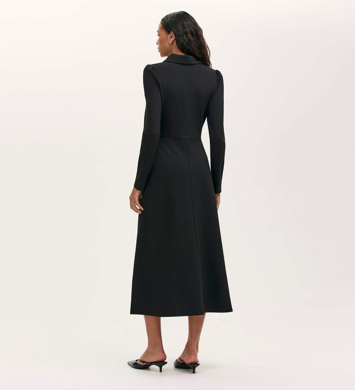 A woman with wavy hair faces away, wearing the Elvira Black Jersey Midi Dress featuring long sleeves, a fitted waist, and flared skirt. She completes the look with black heeled mules against a plain light background.