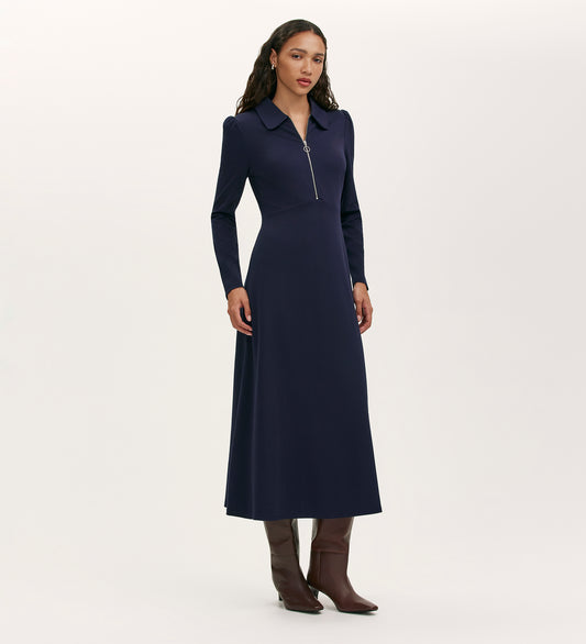 A woman with long wavy hair stands against a plain light background, wearing the Elvira Navy Jersey Midi Dress featuring a collared neckline and front zip, paired with tall brown boots.