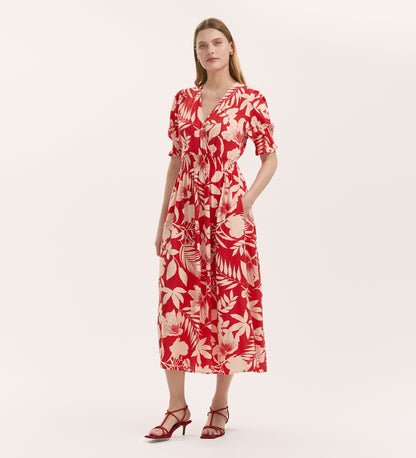 A woman wears the Emily Red Tropical Bloom Midi Dress—featuring a red and white floral print, V-neck, short sleeves, and elasticated waist—paired with red strappy sandals against a plain white background.