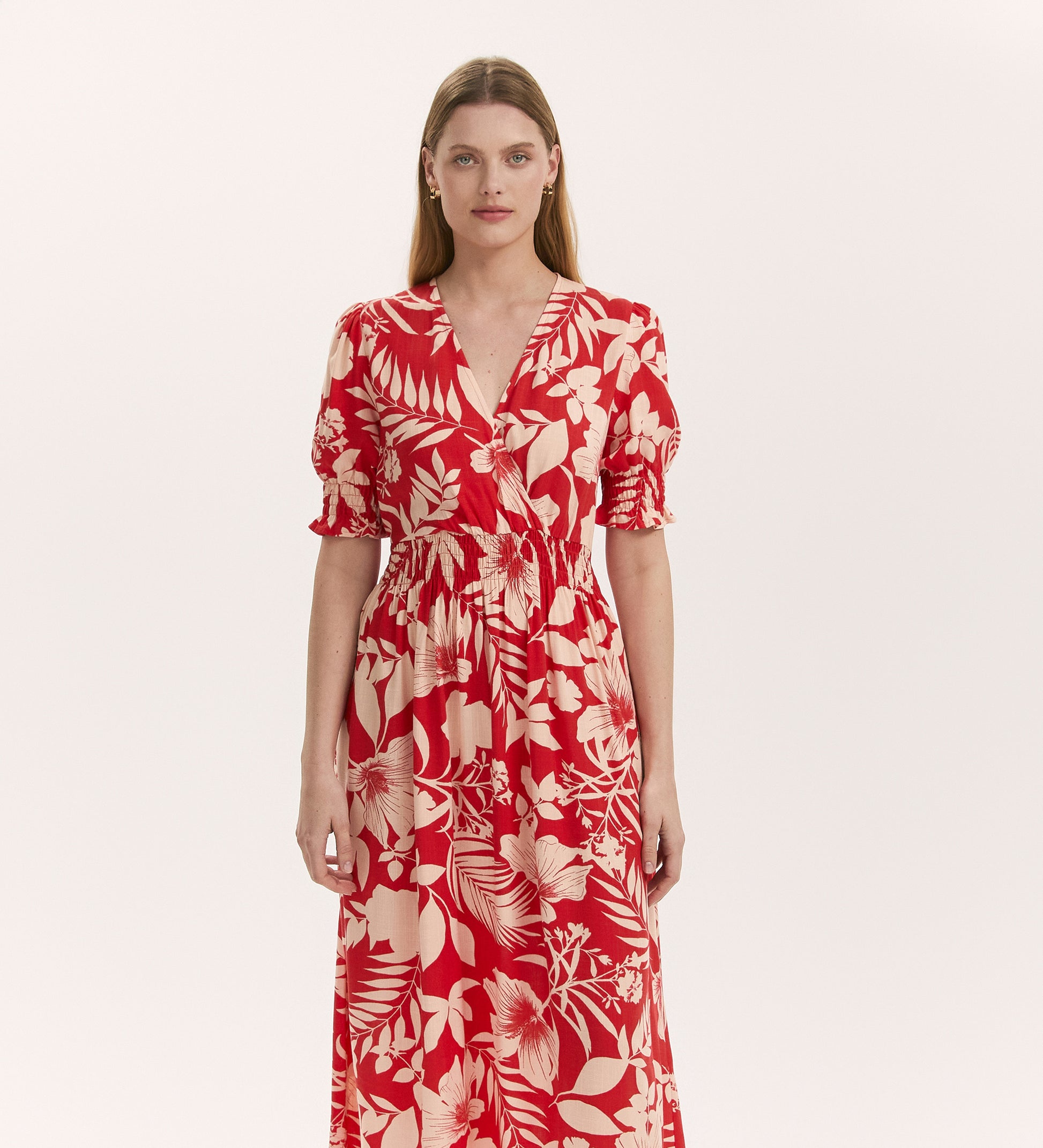 A woman faces forward in the Emily Red Tropical Bloom Midi Dress, featuring a white floral print, short puff sleeves, elastic waist, and v-neckline, set against a plain light background.