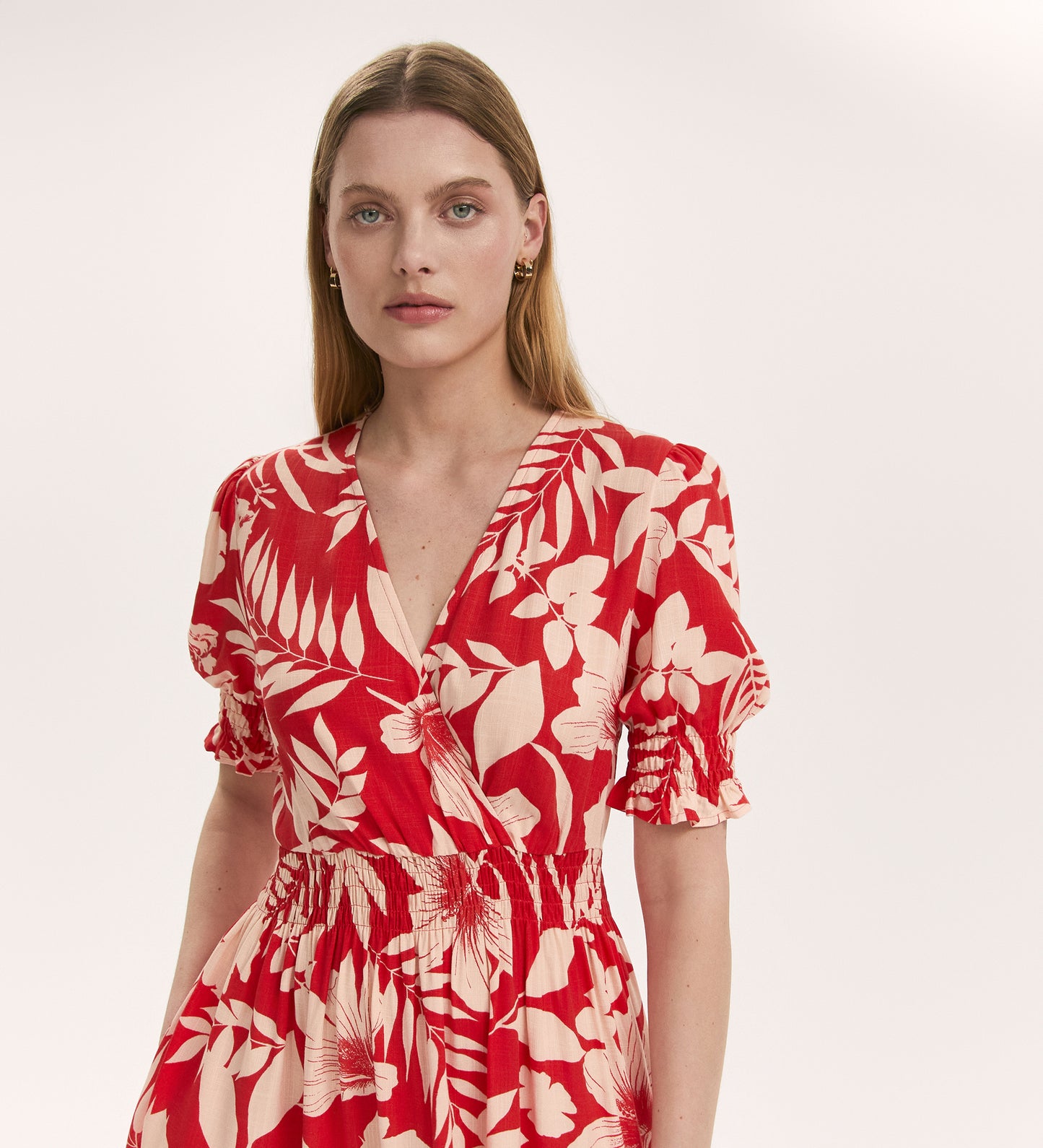 A woman with straight, light brown hair wears the Emily Red Tropical Bloom Midi Dress, featuring a red and cream floral print, short puffed sleeves, elastic waist, and V-neckline. She stands before a plain light background, gazing at the camera.