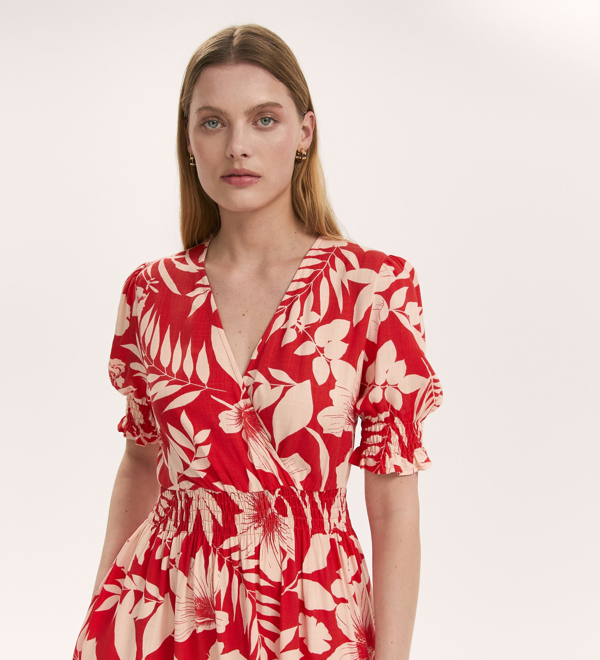 A woman with straight, light brown hair wears the Emily Red Tropical Bloom Midi Dress, featuring a red and cream floral print, short puffed sleeves, elastic waist, and V-neckline. She stands before a plain light background, gazing at the camera.