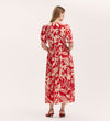 A woman with long blonde hair wears the Emily Red Tropical Bloom Midi Dress featuring a red and white floral print, puff sleeves, and an elasticated waist, styled with strappy red heels, standing against a plain light background facing away.