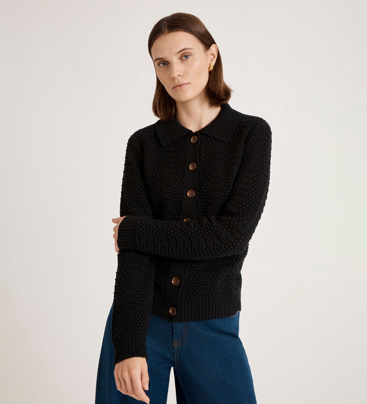 A woman with straight brown hair and a neutral expression stands against a plain light background, wearing blue jeans and the Emlyn Black Knitted Cardigan.