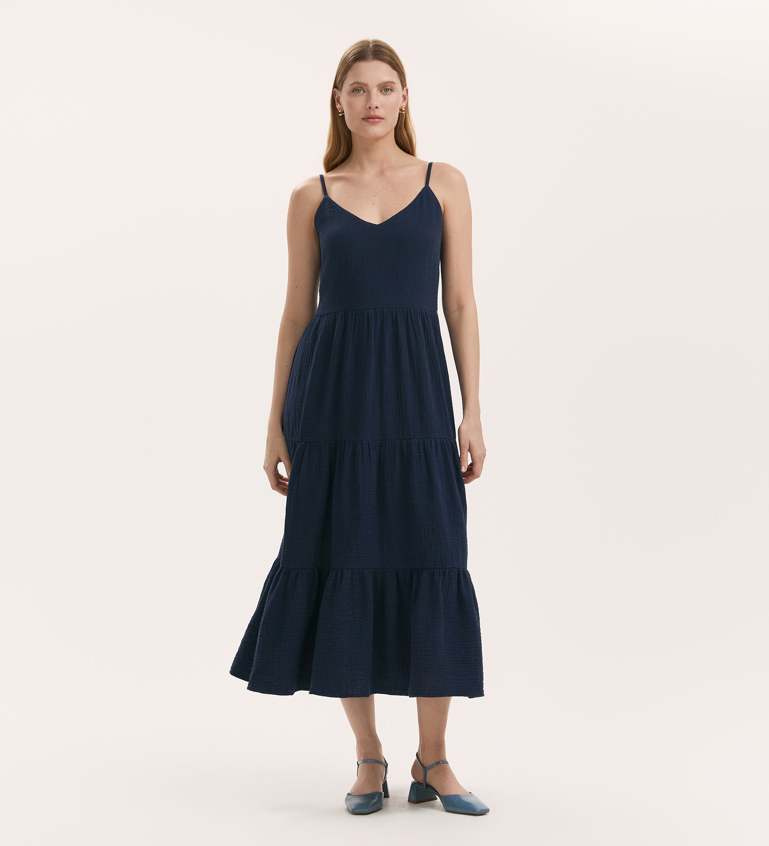 A woman with straight, long hair wears the Emmie Navy Cotton Midi Tiered Dress, featuring thin straps, a V-neckline, and a tiered skirt. She pairs it with blue strap-heeled shoes. The background is plain and light.