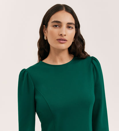 A woman with long dark wavy hair wears the Eva Bottle Green Ponte Jersey Short Dress, featuring a fitted mini silhouette with subtle puffed shoulders and princess seams. She pairs it with gold hoop earrings and stands against a plain light background.