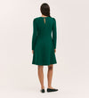 A woman with dark hair in a loose braid wears the Eva Bottle Green Ponte Jersey Short Dress, featuring princess seams and a back keyhole opening. She pairs it with black flats and stands against a plain light background, facing away from the camera.