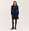 A woman with long brown hair and a neutral expression wears the Eva Navy Ponte Jersey Short Dress against a plain light background, paired with black tights and ankle boots.