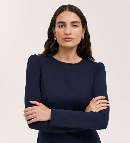 A woman with long wavy brown hair stands confidently against a plain light background, wearing the Eva Navy Ponte Jersey Short Dress featuring princess seams.