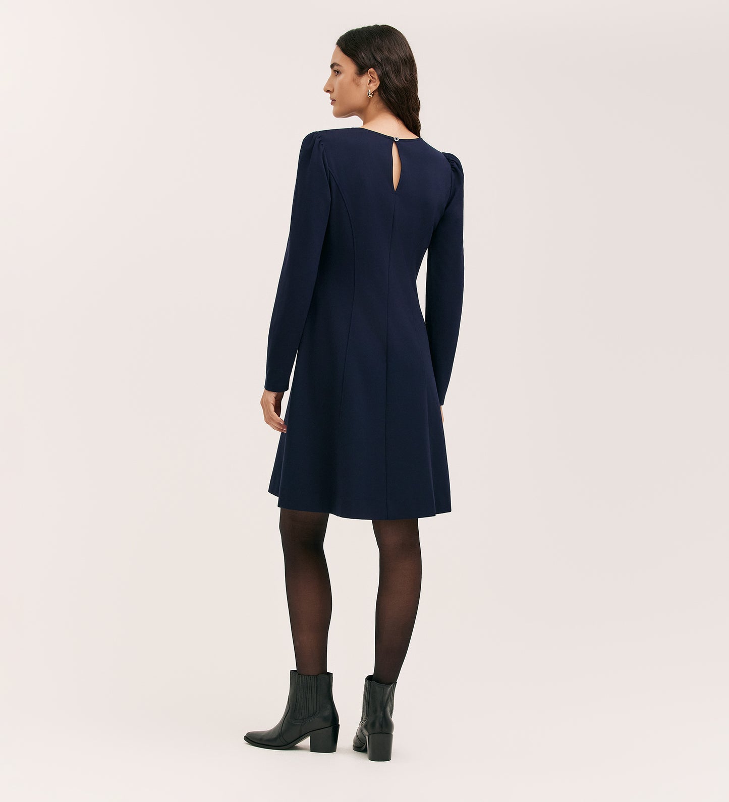 A woman faces away, wearing the Eva Navy Ponte Jersey Short Dress with long sleeves, princess seams, and a back keyhole detail, paired with black tights and ankle boots against a plain light background.