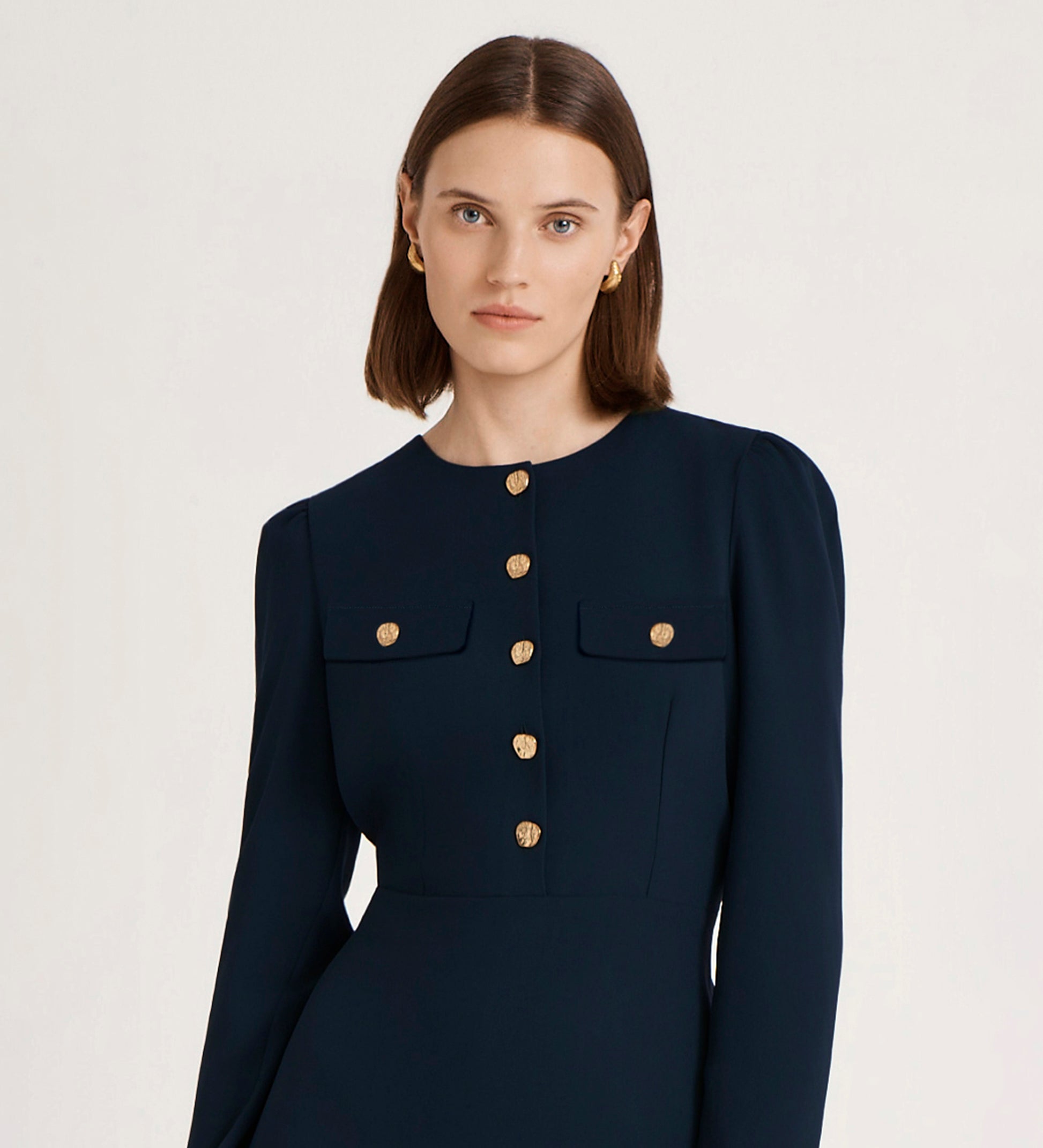 A woman with straight brown hair, wearing gold earrings and the Evie Navy Crepe Short Dress featuring gold buttons and chest pockets, stands against a plain light background.