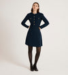 A woman with straight, shoulder-length brown hair stands against a plain light background, wearing the Evie Navy Crepe Short Dress featuring gold button detailing, paired with black tights and shoes. She maintains a neutral expression.