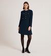 A woman with straight brown hair and a neutral expression wears the Evie Navy Crepe Short Dress, featuring gold button details, black tights, and black shoes, standing against a plain light background.