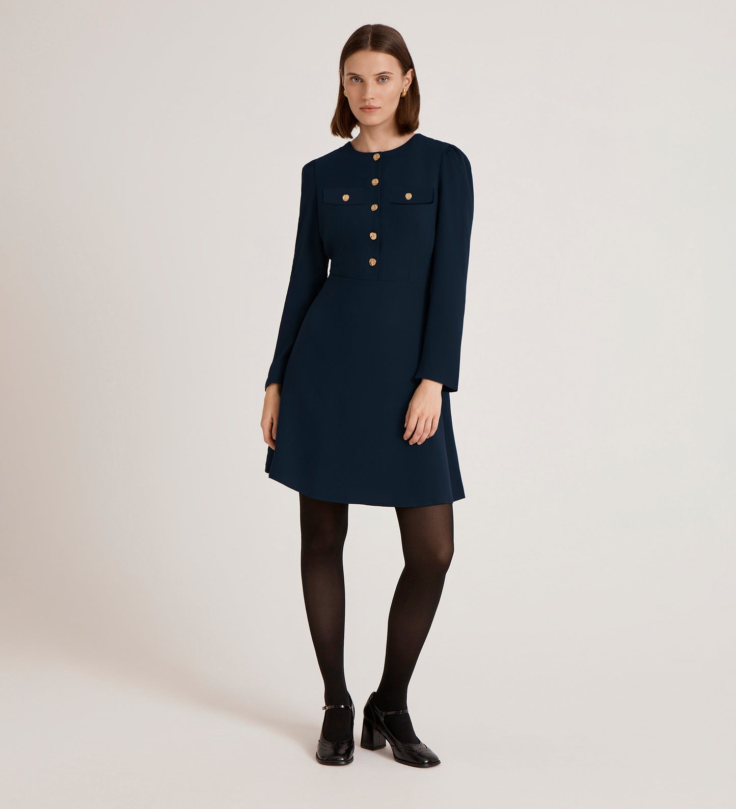 A woman with straight brown hair and a neutral expression wears the Evie Navy Crepe Short Dress, featuring gold button details, black tights, and black shoes, standing against a plain light background.