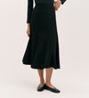 A person stands against a plain light background wearing the Fabiola Black Crepe Midi Skirt with a long-sleeved black top and black flats. Their head is not visible in the image.