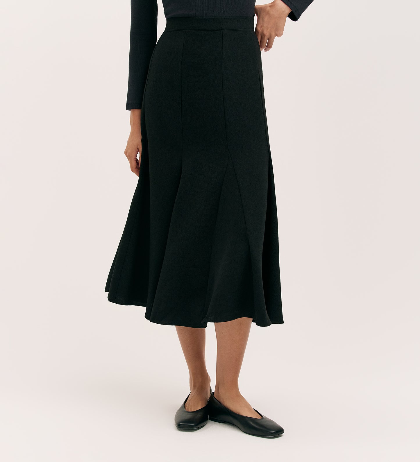A person stands against a plain light background wearing the Fabiola Black Crepe Midi Skirt with a long-sleeved black top and black flats. Their head is not visible in the image.