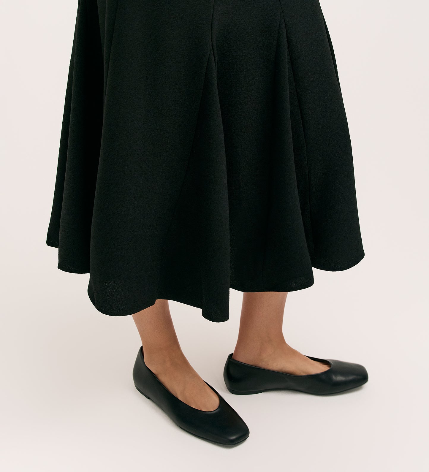 A person models the Fabiola Black Crepe Midi Skirt with black flats, standing against a plain, light background. Only their lower legs and feet are shown.
