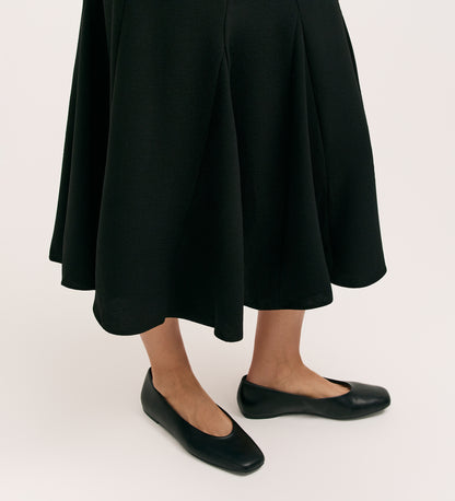 A person models the Fabiola Black Crepe Midi Skirt with black flats, standing against a plain, light background. Only their lower legs and feet are shown.