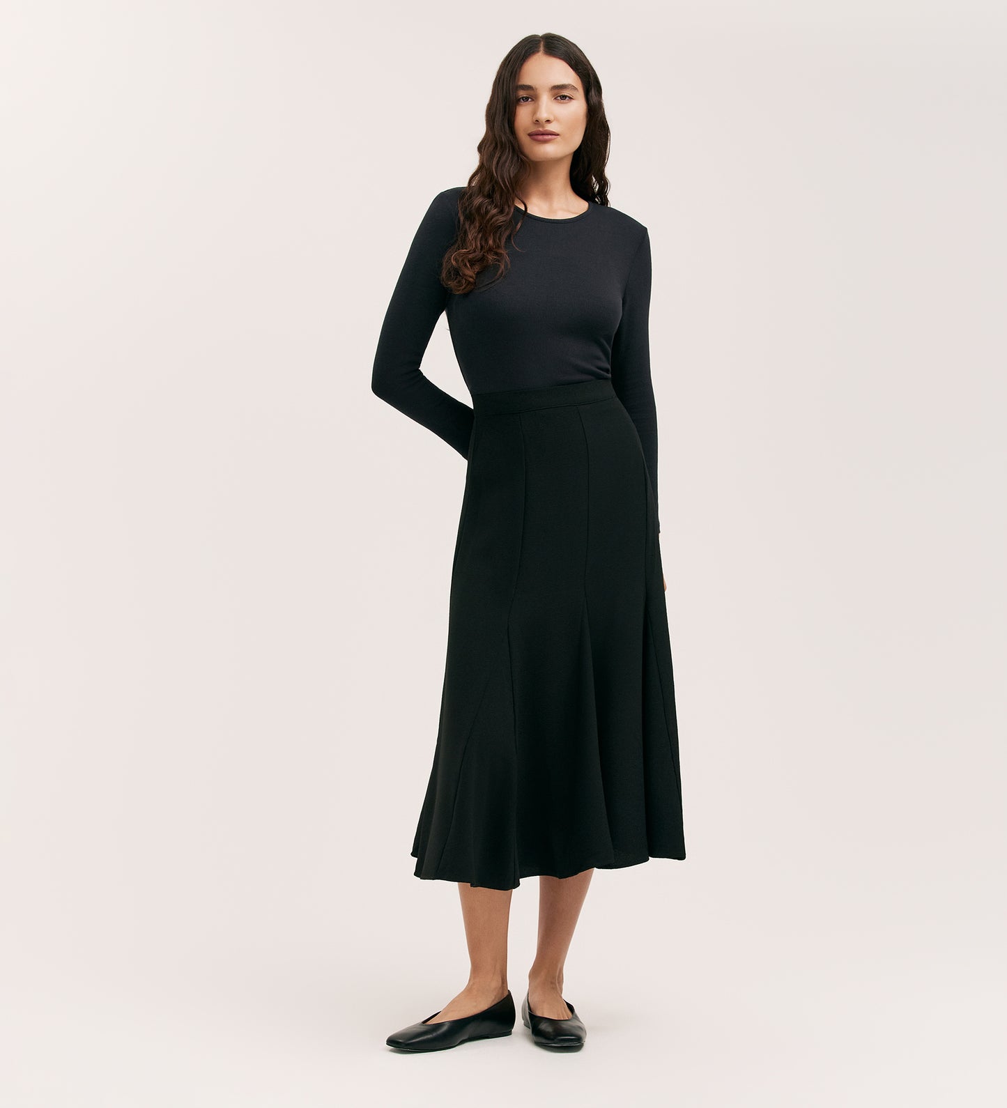 A woman with long, wavy dark hair wears a fitted, long-sleeve black top and the Fabiola Black Crepe Midi Skirt, paired with classic black flats. She stands against a plain light background.