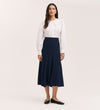 A woman with long dark hair stands against a plain background, wearing a white blouse and the Fabiola Navy Crepe Midi Skirt, paired with black flats. She gazes ahead with a neutral expression.