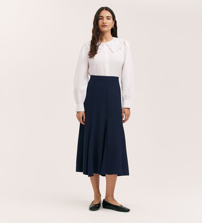 A woman with long dark hair stands against a plain background, wearing a white blouse and the Fabiola Navy Crepe Midi Skirt, paired with black flats. She gazes ahead with a neutral expression.