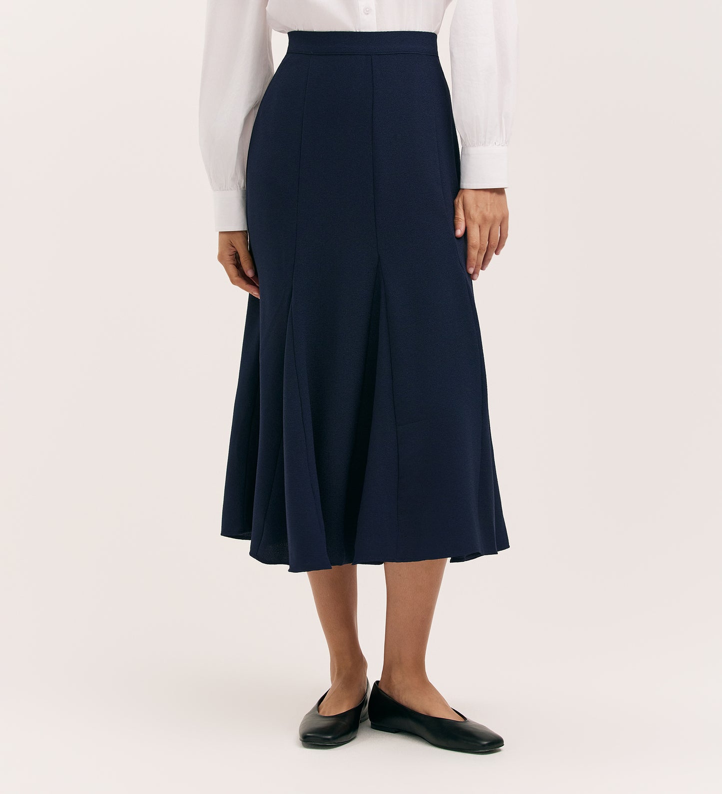 A person wearing a white long-sleeve blouse, the Fabiola Navy Crepe Midi Skirt, and black flat shoes stands against a plain light background.