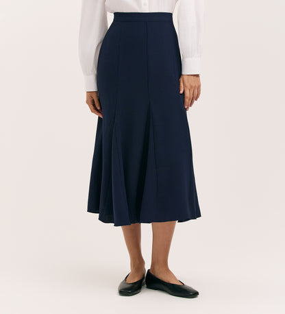 A person wearing a white long-sleeve blouse, the Fabiola Navy Crepe Midi Skirt, and black flat shoes stands against a plain light background.