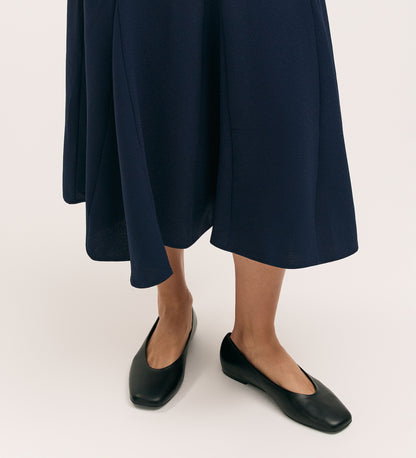 A person stands on a light surface, wearing the Fabiola Navy Crepe Midi Skirt and black square-toe flats; only their lower legs and feet are visible.