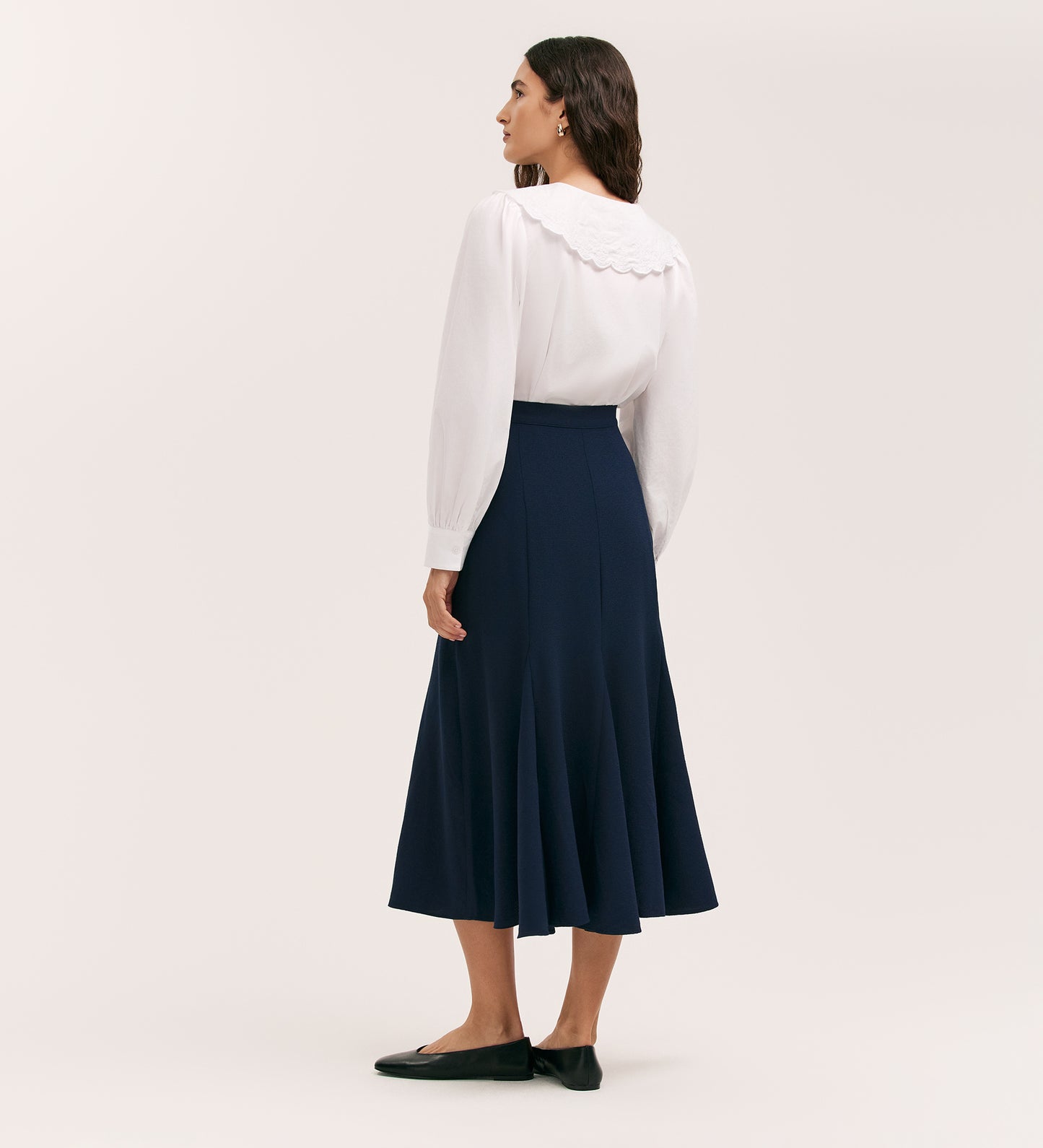 A woman with medium-length dark hair stands facing away in a white long-sleeve blouse and the Fabiola Navy Crepe Midi Skirt, paired with black flats. The background is plain and light-colored.