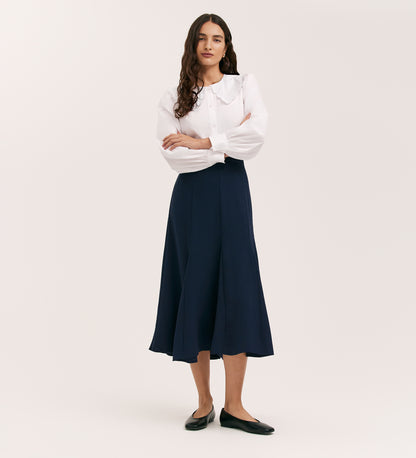 A woman with long, wavy hair stands against a plain background wearing the Fabiola Navy Crepe Midi Skirt, a white blouse with a wide collar, and black flats. Her arms are crossed.