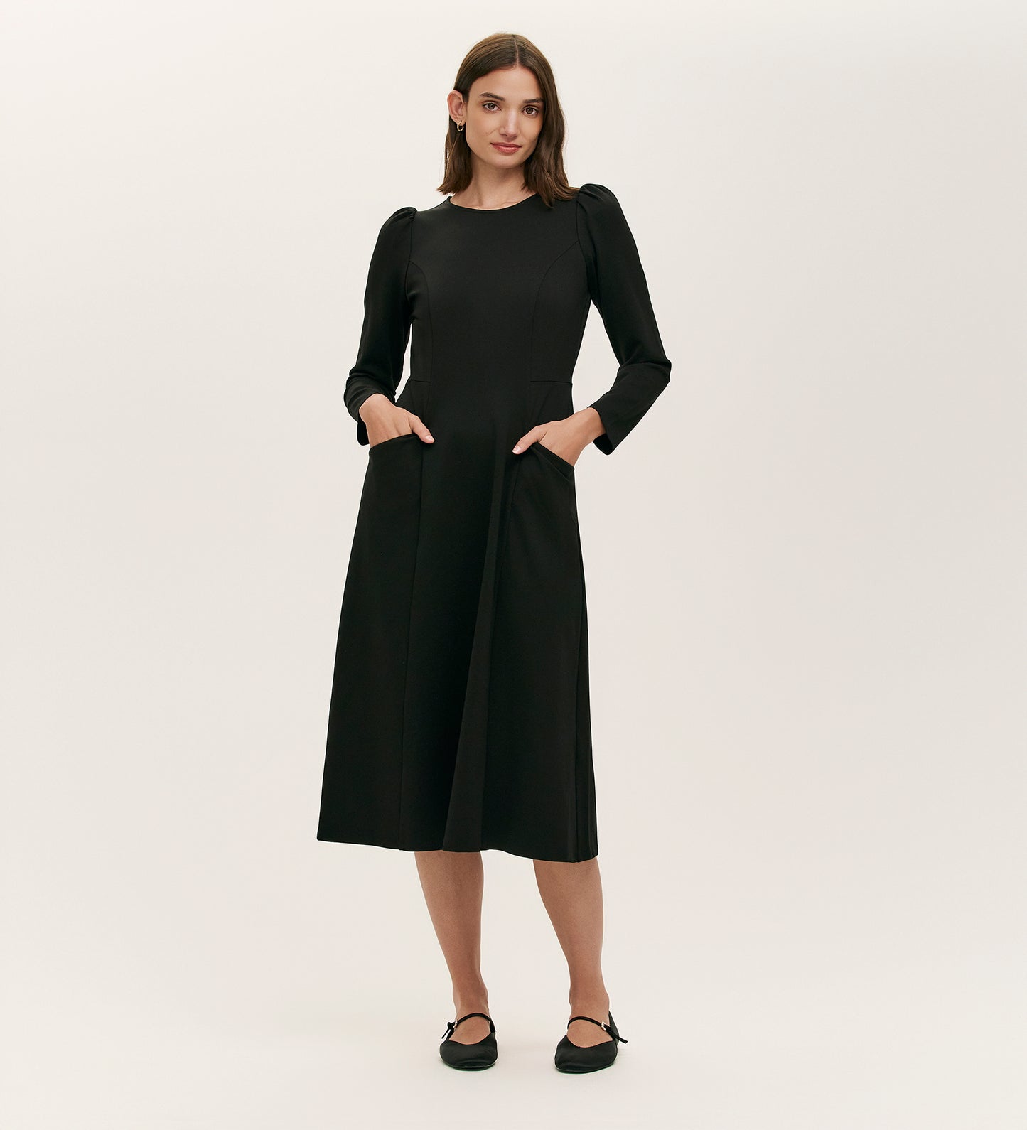 A woman with straight brown hair stands against a plain background wearing the Felice Black Ponte Jersey Midi Dress, featuring long sleeves, subtle puff shoulders, pockets, and a tailored silhouette. She completes the look with black flat shoes.