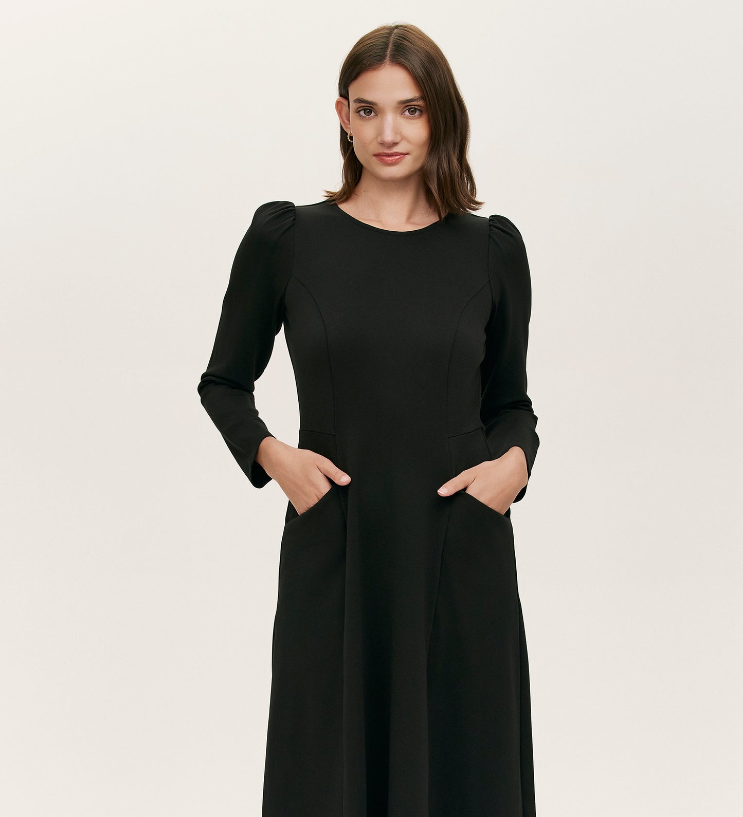 A woman with medium-length brown hair wears the Felice Black Ponte Jersey Midi Dress, featuring long sleeves, subtle puffed shoulders, and a tailored silhouette, standing with hands in pockets against a plain light background.