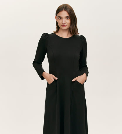 A woman with medium-length brown hair wears the Felice Black Ponte Jersey Midi Dress, featuring long sleeves, subtle puffed shoulders, and a tailored silhouette, standing with hands in pockets against a plain light background.