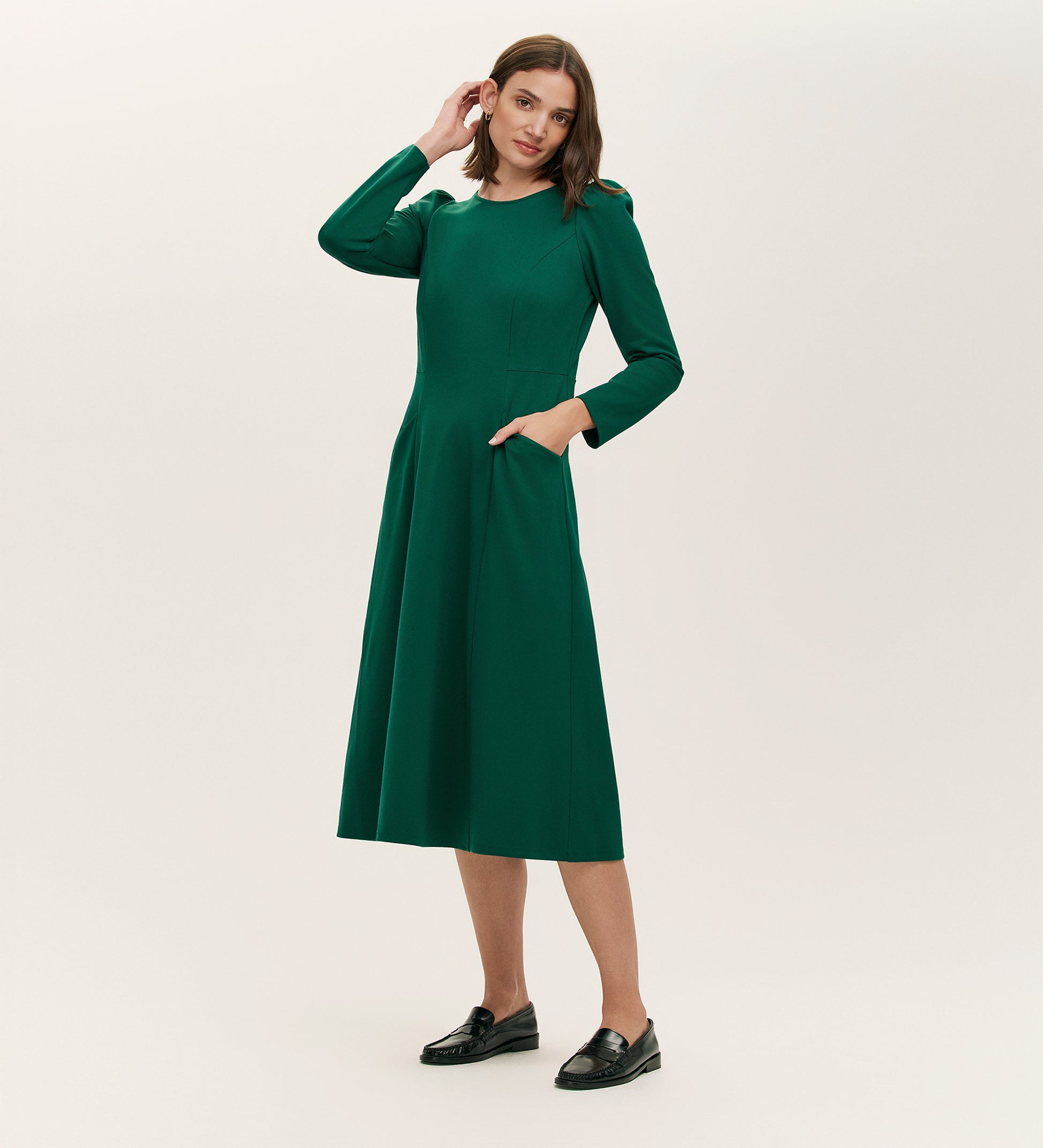 A woman with straight brown hair stands against a light background, wearing the Felice Bottle Green Ponte Jersey Midi Dress with pockets and black loafers. She touches her head with one hand and gazes slightly to the side.