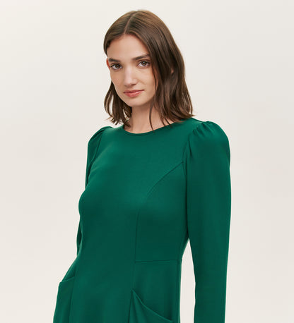 A woman with shoulder-length brown hair wears the Felice Bottle Green Ponte Jersey Midi Dress, featuring long sleeves, subtle puffed shoulders, and a tailored silhouette, standing against a plain light background.