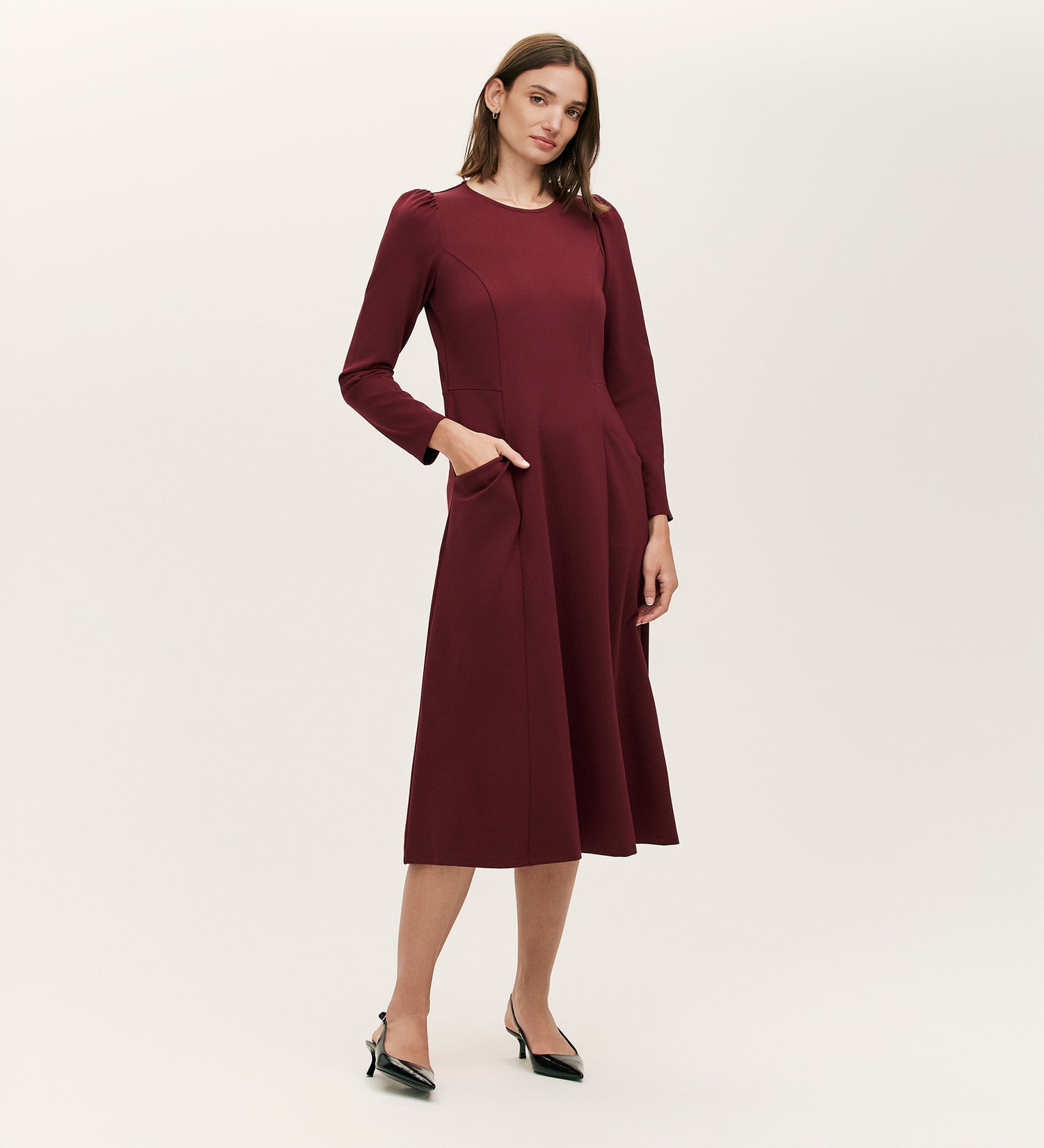 A woman stands against a plain light background, wearing the Felice Burgundy Ponte Jersey Midi Dress with a tailored silhouette, long sleeves, practical pockets, and black pointed-toe heels.
