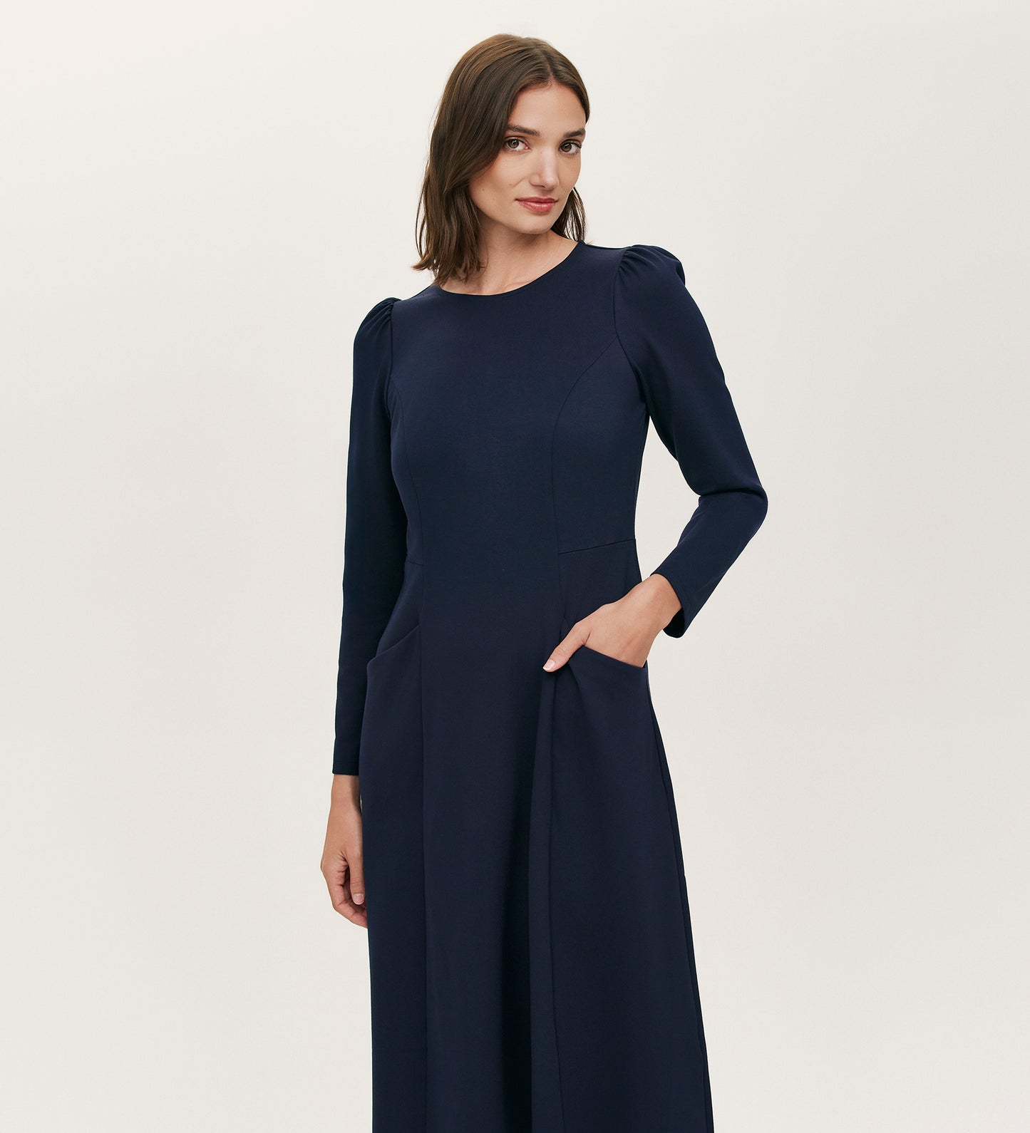 A woman with straight brown hair wears the Felice Navy Ponte Jersey Midi Dress, which features subtle puffed shoulders, a tailored silhouette, and pockets, standing against a plain light background.