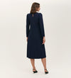 A woman with shoulder-length brown hair and black heels faces away, wearing the Felice Navy Ponte Jersey Midi Dress with a tailored silhouette and buttoned keyhole back against a plain light background.
