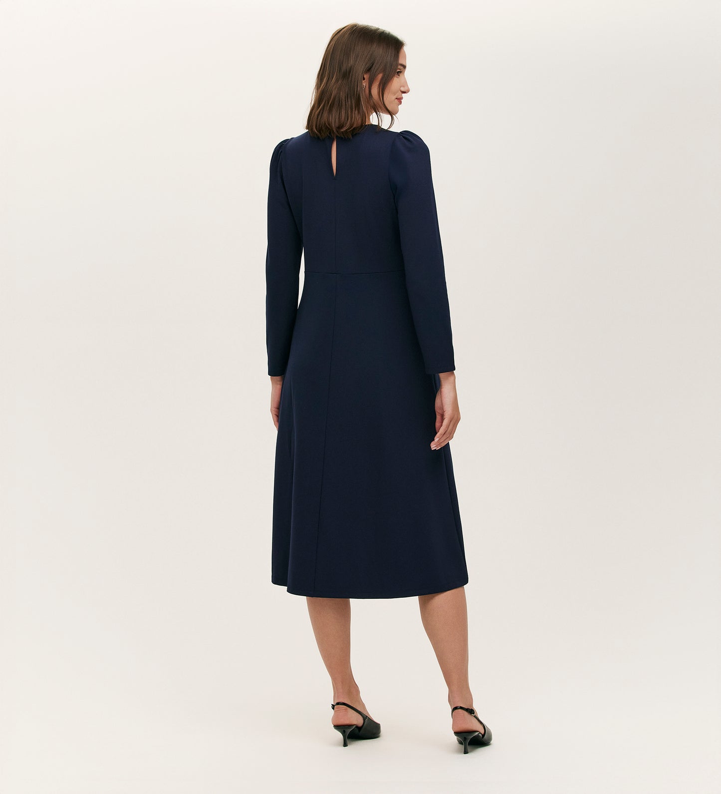 A woman with shoulder-length brown hair and black heels faces away, wearing the Felice Navy Ponte Jersey Midi Dress with a tailored silhouette and buttoned keyhole back against a plain light background.