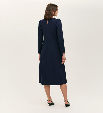 A woman with shoulder-length brown hair and black heels faces away, wearing the Felice Navy Ponte Jersey Midi Dress with a tailored silhouette and buttoned keyhole back against a plain light background.