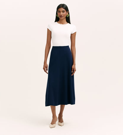 A woman with long dark hair and a neutral expression wears a fitted white short-sleeve top, the Mae Navy Ponte Jersey Midi Skirt with an elasticated waistband, and beige pointed-toe heels against a plain background.