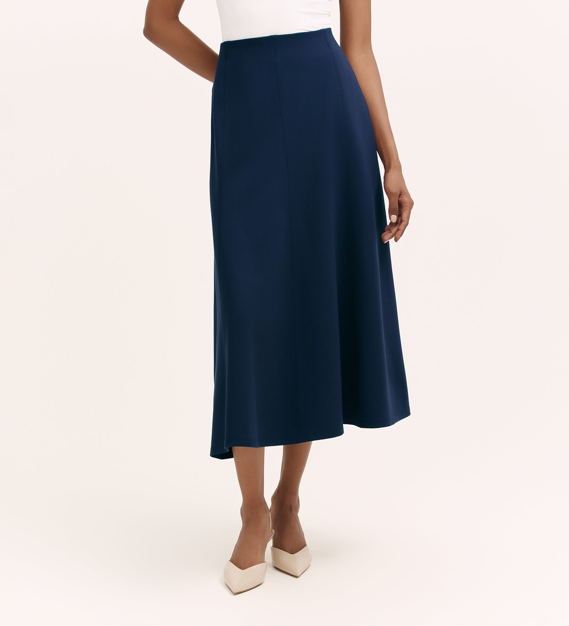 A woman wears a white sleeveless top paired with the Mae Navy Ponte Jersey Midi Skirt, featuring an elasticated waistband, and pointed-toe beige heels. She stands against a plain light background with one arm relaxed by her side.