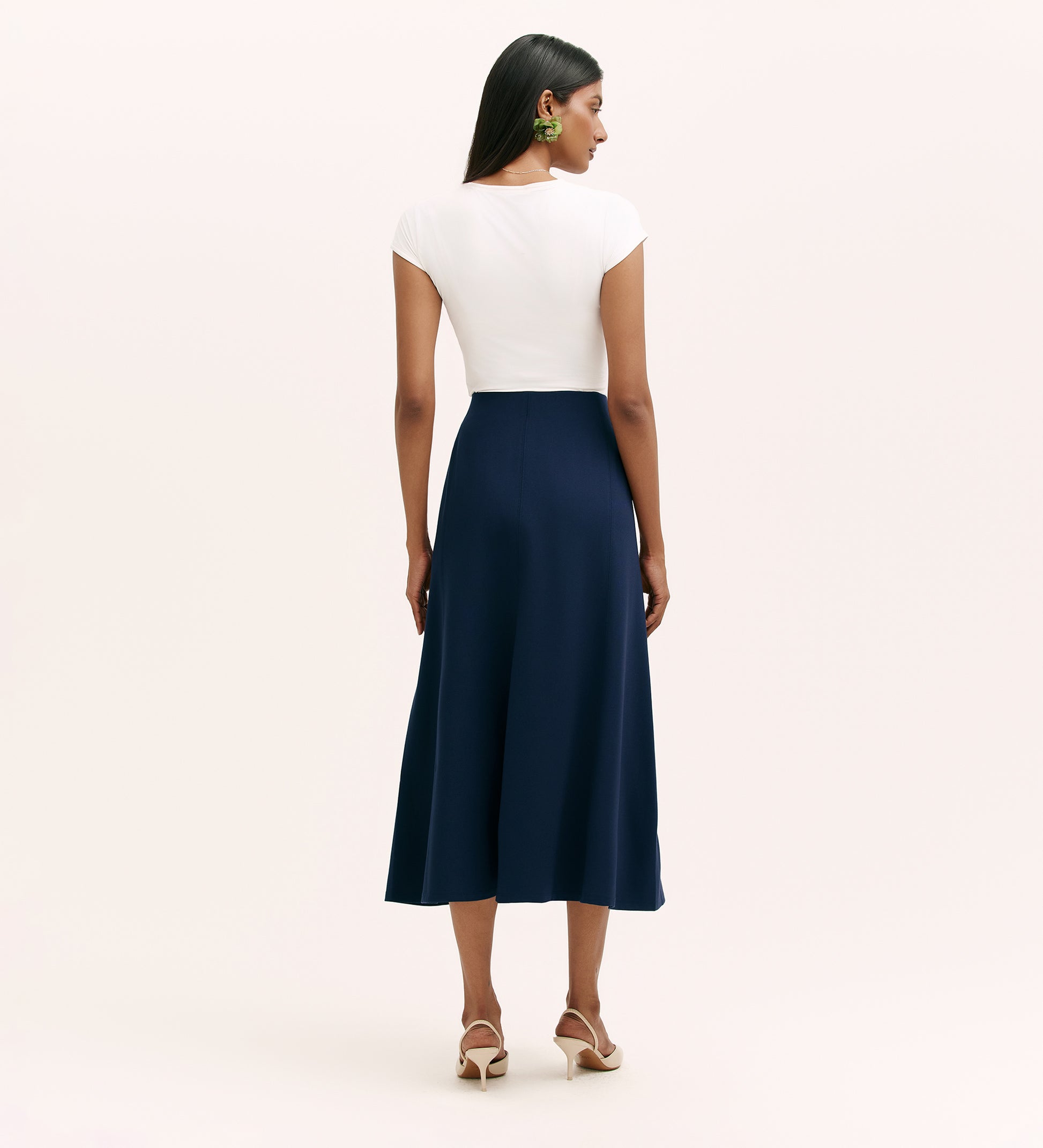 A woman faces away, wearing a white fitted cap sleeve top, the Mae Navy Ponte Jersey Midi Skirt with an elasticated waistband, beige heeled sandals, and green statement earrings against a plain light background.