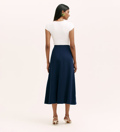 A woman faces away, wearing a white fitted cap sleeve top, the Mae Navy Ponte Jersey Midi Skirt with an elasticated waistband, beige heeled sandals, and green statement earrings against a plain light background.