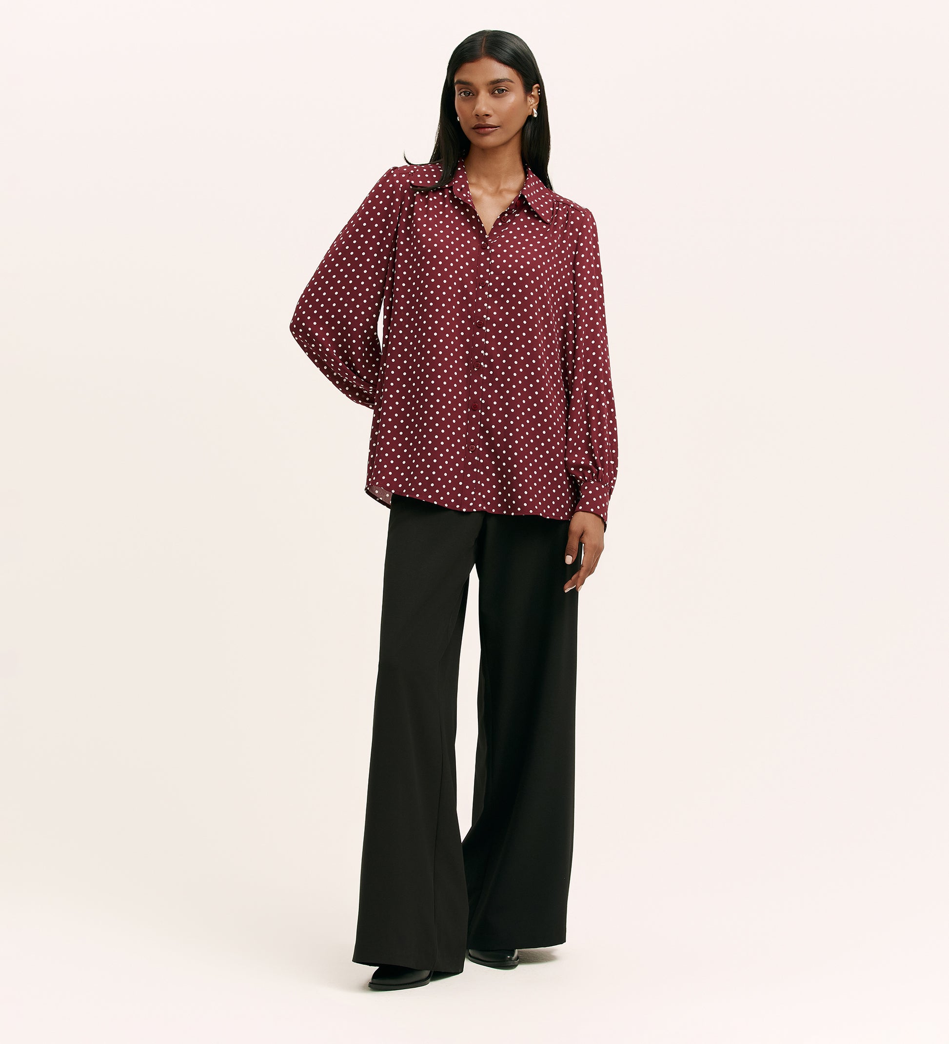 A woman stands against a plain background wearing the Melina Burgundy Spot Crepe Blouse—a relaxed fit, lightweight crepe shirt with white polka dots and a button-through front—paired with black wide-leg trousers. She poses with a relaxed expression.