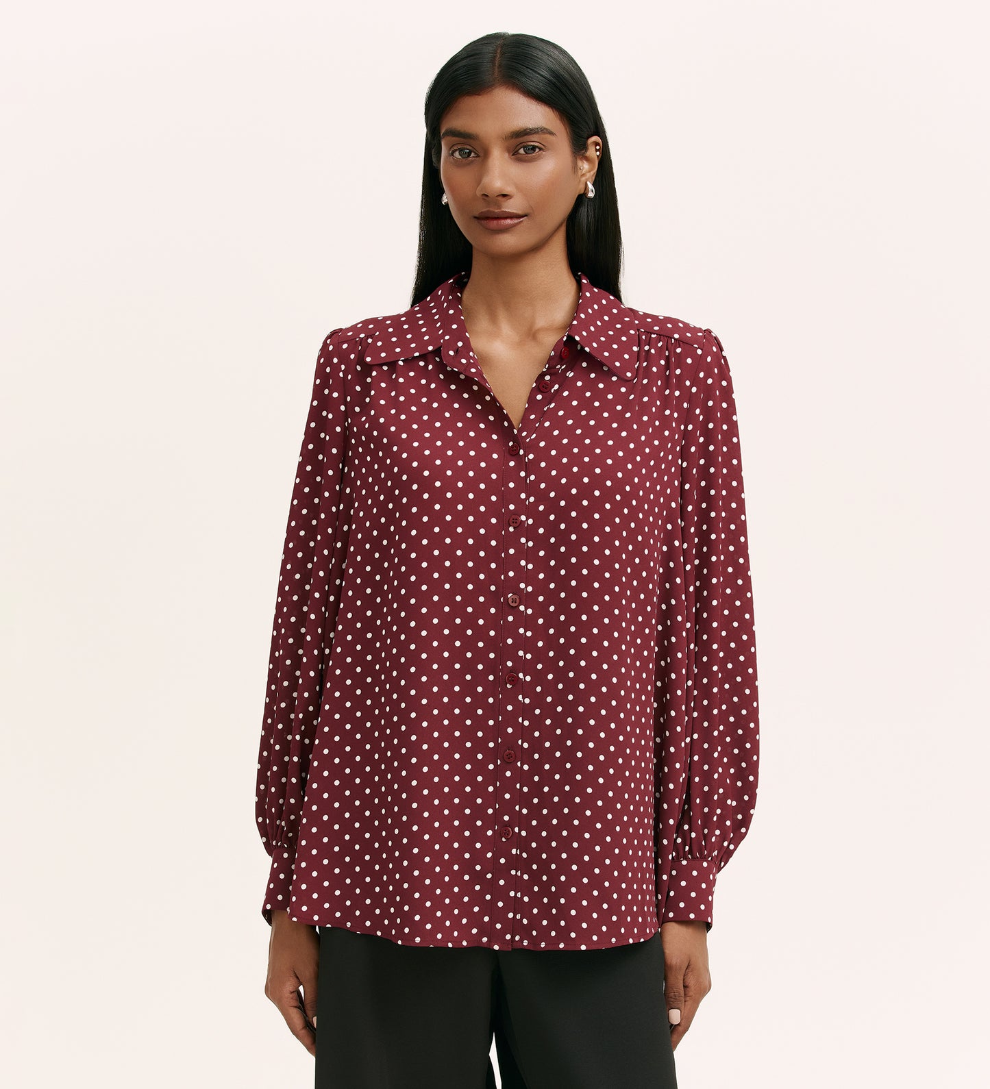 A woman with long dark hair wears the Melina Burgundy Spot Crepe Blouse, a maroon button-up with white polka dots and long sleeves, in a relaxed shirt style, paired with black pants against a plain light background.