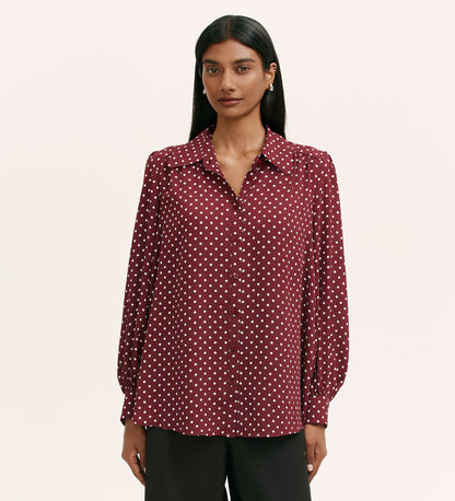 A woman with long dark hair wears the Melina Burgundy Spot Crepe Blouse, a maroon button-up with white polka dots and long sleeves, in a relaxed shirt style, paired with black pants against a plain light background.
