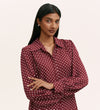 A woman with long, straight dark hair wears the Melina Burgundy Spot Crepe Blouse, featuring white polka dots, a button-through front, long sleeves, and a relaxed fit. She stands against a plain light background with her arms crossed.