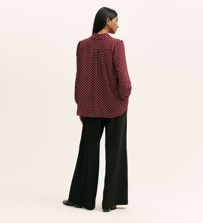 A woman with long, straight dark hair stands against a light background wearing the Melina Burgundy Spot Crepe Blouse—a relaxed fit maroon shirt with white polka dots—paired with loose black trousers.