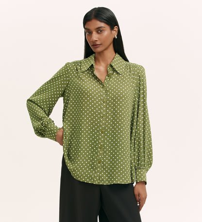 A woman with long dark hair wears the Melina Green Spot Chiffon Blouse with white polka dots and black pants, standing with one hand on her hip against a light background.